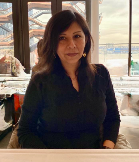 A brunette haired person wearing a dark black blouse with gold dangling earrings.